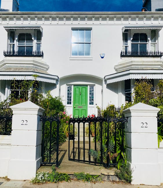 A white, two-story residential building with decorative black wrought iron balcony railings and bay windows. The front garden features a small paved pathway leading to a bright green front door, flank